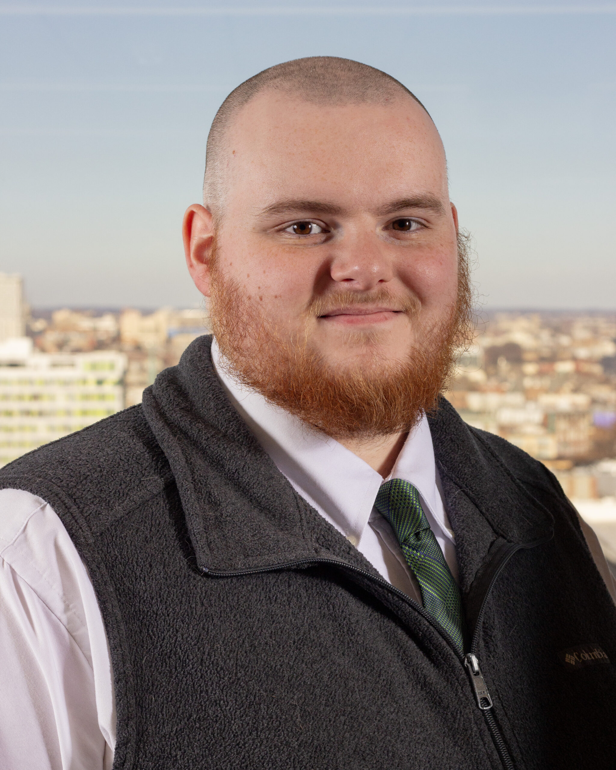Cole Lyons outdoors above a city skyline, wearing a white shirt, tie, and dark vest